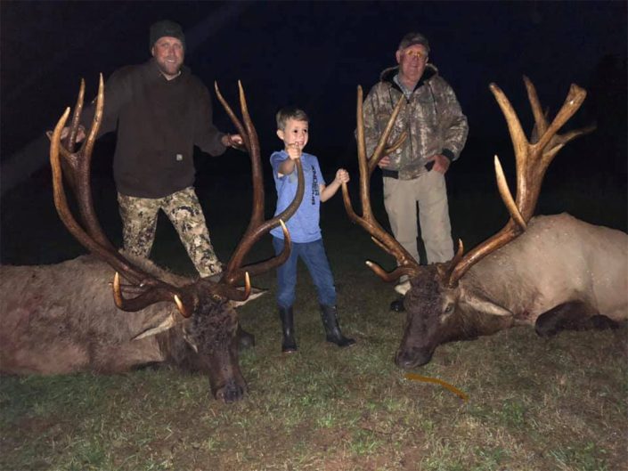 two men and a child pose with a pair of trophy elk kills 